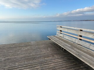 A wooden bench on a pier overlooking a calm lake under a partly cloudy sky. The tranquil water and soft clouds create a peaceful and relaxing atmosphere. Ideal for concepts of solitude and serenity.
