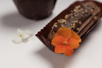 Chocolate ?clair in a brown wrapper with a smooth chocolate glaze and an orange edible flower on top, with a blurred background.