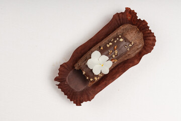 A close-up of a chocolate ?clair in a brown wrapper, decorated with a white edible flower and...