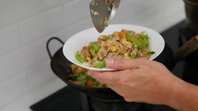 Close-up of a person serving freshly cooked fried rice with vegetables, including green beans and red bell peppers, from a wok onto a white plate using a metal spatula in a home kitchen setting