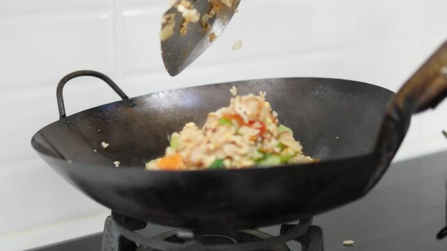 Close-up of a wok on a gas stove with freshly cooked fried rice being scooped up with a metal spatula, featuring vegetables like green bell peppers and carrots in a home kitchen