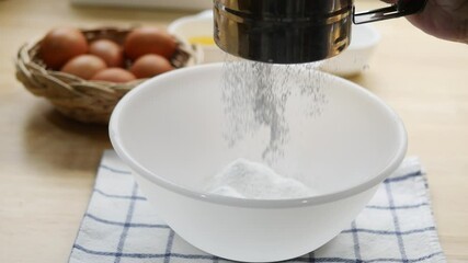Here is a 200-character image title for your image:

Close-up of a person sifting flour into a white mixing bowl while preparing ingredients for baking. A basket of fresh brown eggs and a small bowl