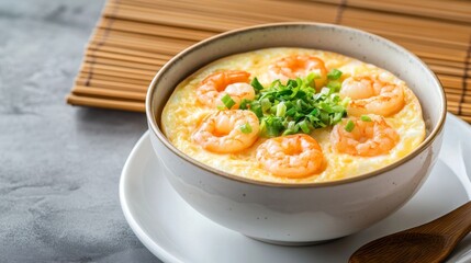 A minimalist shot of a bowl of steamed egg, topped with shrimp and green onions, set on a white plate with a wooden spoon and a bamboo mat in the background.