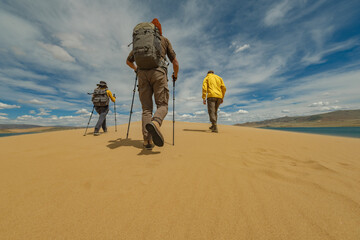 A group of tourists in bright clothes with backpacks on their backs walk along a sand dune against the background of a lake and sky. People are walking on the sand.