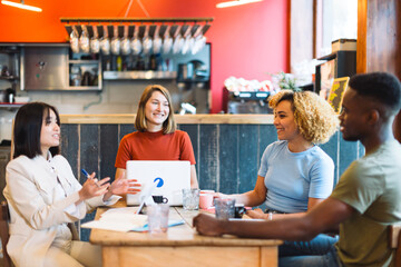 Four young professionals discussing ideas in a casual cafe setting with laptops, documents, and drinks