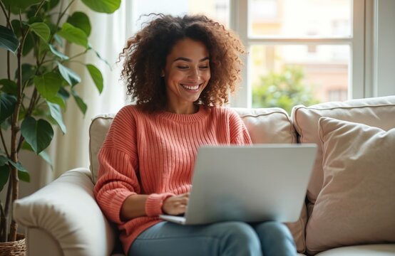Young woman sits on sofa using laptop. She is happy surfing web, shopping online, watching video or elearning. Girl looking at computer ordering products on website.