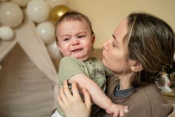 Portrait of a crying baby boy. The child is teething, he is naughty and asks for his mother hands. Mom is calming the baby.