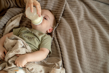 Happy baby boy is drinking milk formula from a bottle lying on the bed. Kid aged 1 year old