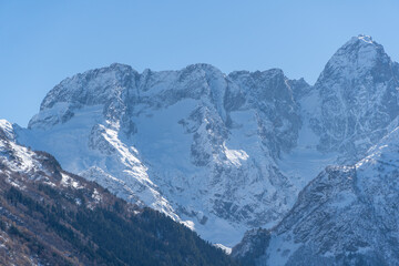 Fototapeta premium Snowy peaks and glaciers cover giant rocks. Pine and spruce forests grow at foot of mountains. which Majestic mountains against blue sky. Sunny day. Autumn 2024. Karachay-Cherkessia. Dombay.