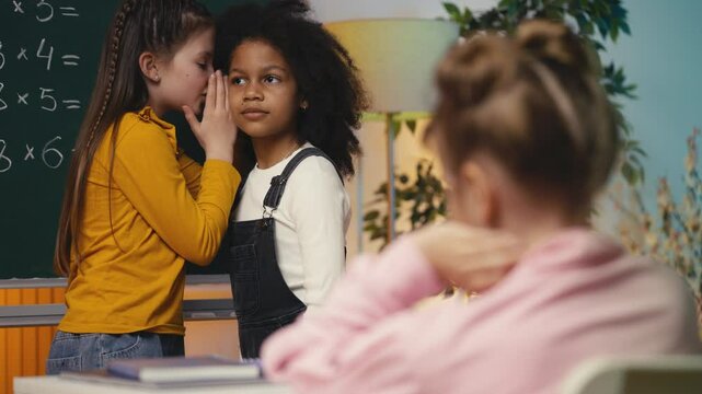 Two multiethnic schoolgirls gossiping about their lonely classmate in the class