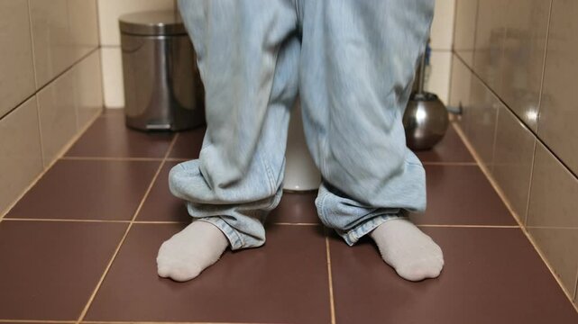 A man sits on the toilet in a bathroom with his pants down, after successfully finishing the process, his legs dance with relief. He then pulls up his pants, flushes the toilet. Constipation relief