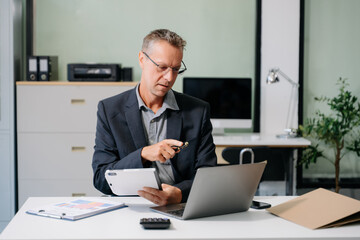 Confident businessman working on finance analysis with a tablet and laptop at office desk.