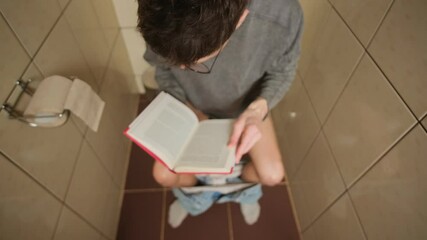 Young man sitting on the toilet with his pants down, engrossed in reading a book, viewed above. He wears glasses and a gray sweater, fully absorbed in the text, creating a moment concentration.
