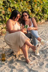 Two women enjoying wine and using smartphone on beach