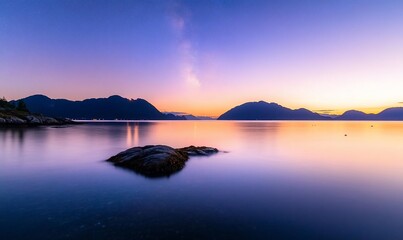 Naklejka premium Serene sunset over calm water with silhouetted mountains and a lone rock.