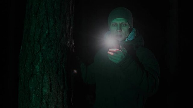 Lost man emerging from darkness in a forest at night, using a phone flashlight to navigate, stopping by a tree to check his map, depicting the concept of being lost in the woods.