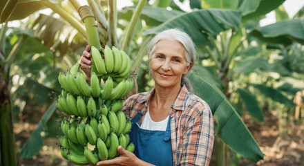 Elderly caucasian female farmer holding green bananas in tropical plantation.