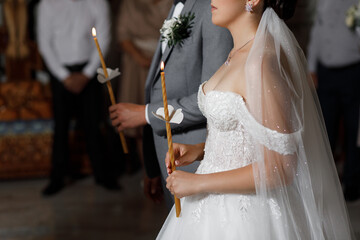 The newlyweds stand nearby, holding lit candles in their hands. Woman in a white wedding dress with...