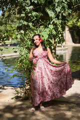 Fototapeta premium Moroccan woman, young, beautiful, brunette, in cashmere dress, with a hibiscus flower in her ear surrounded by green leaves, next to a lake. Concept ethnicity, diversity, identity. Vertical position.