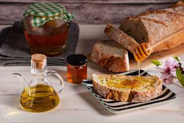 Toasted slice of bread with olive oil and honey, on a white table with an oil dispenser, a jar of honey, and a loaf of bread sliced, close-up.