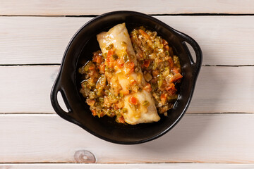 Hake loin with sautéed vegetables on a black ceramic plate, on a white wooden table, top view.