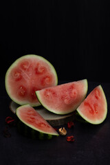 Vertical image of freshly harvested watermelon cut into several pieces on a rustic wooden log on a dark background. Tropical fruit still life