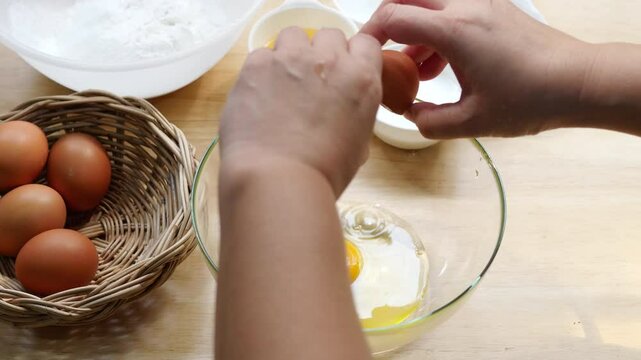 Hands Cracking Egg into Glass Bowl on Wooden Kitchen Counter with Basket of Eggs and Baking Ingredients in Background