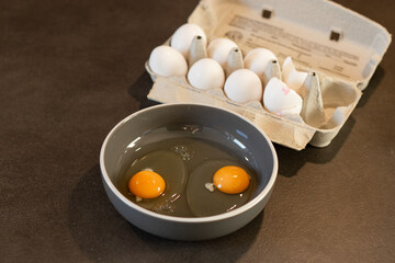 Close up of two cracked eggs in a bowl on kitchen table with dozen of white eggs in a cardboard