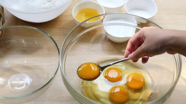 Cracking and Separating Eggs in Glass Bowl for Baking &ndash; Preparing Cake Batter with Flour, Sugar, and Butter on Wooden Kitchen Counter with Ingredients in Small Bowls for Homemade Dessert