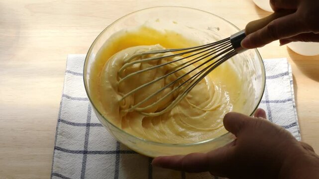 Hand Mixing Smooth Cake Batter with Whisk in Glass Bowl &ndash; Baking Process on Wooden Kitchen Counter with Checkered Cloth, Preparing Homemade Dessert with Eggs, Flour, and Sugar