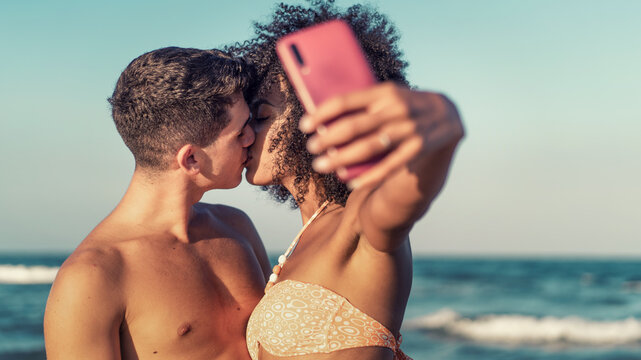 Interracial couple kissing while taking a selfie at the beach - inclusive summer love