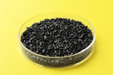 Black plastic granules in petri dish on yellow background, closeup