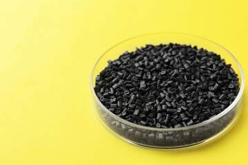 Black plastic granules in petri dish on yellow background, closeup. Space for text