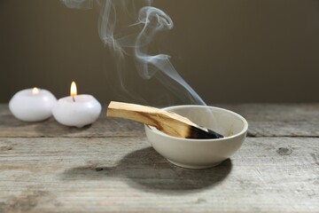 Smoldering palo santo stick and burning candles on wooden table against grey background, closeup