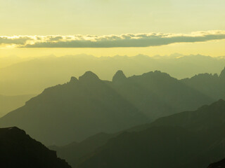 Dolomites mountains under golden sky aerial view, peaks fading into mist, creating magical layered effect. Soft sunlight enhances rugged terrain, bringing depth to breathtaking landscape. Travel