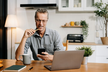 Lifestyle people, using laptop computer and smartphone sitting in the kitchen. Happy mature male,...