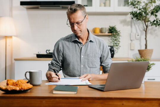 Mature male entrepreneur working online from home, managing documents and laptop at kitchen desk.  modern small business owner embracing remote work.