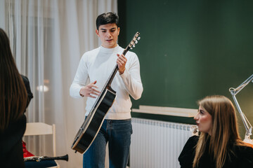 A young man in a white shirt plays a guitar as others observe attentively in a casual indoor environment. The setting conveys creativity, interaction, and focus, promoting artistic expression.