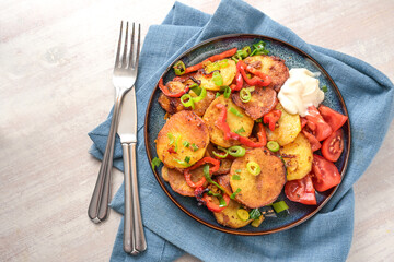 Fried potato slices, red bell pepper and green spring onions in a plate on a blue napkin and cutlery on a light painted wooden table, top view from above, copy space