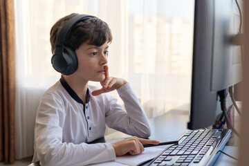 A child aged 7-9 sits at home behind a computer, writing assignments in a notebook. The image captures remote learning, digital study routines,  challenges associated with autism and ADHD. copyspace