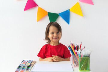 Smiling little girl in a red T-shirt sitting at a table with art supplies, ready to create....