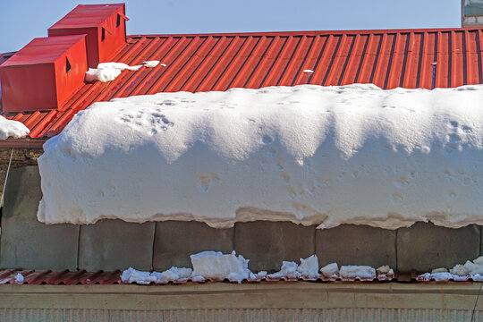 Thick snow on red metal roof melting under sun, creating risk of collapse and falling icicles.