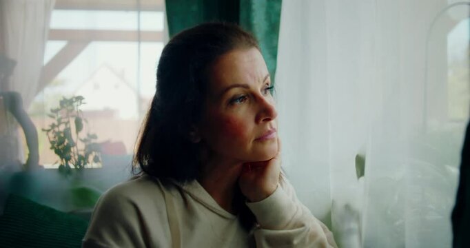 Close-up of a woman looking outside with a calm and thoughtful expression, resting her chin on her hand, framed by soft curtains and a cozy indoor setting