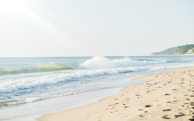 Golden sandy beach with ocean waves crashing under the warm sunlight. Footprints in the sand lead towards the distant coastline with green hills and small houses. A peaceful and scenic seaside view.
