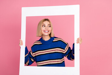 Happy young woman in a stylish striped blue sweater holding a white frame against a pink background