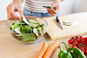 Young woman preparing healthy salad in a modern kitchen, adding fresh cucumber into bowl with lettuce. Healthy lifestyle and food concept