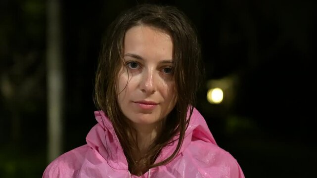 Young woman in bright pink rain jacket at night. Night portrait featuring girl in vibrant pink hooded raincoat. Pink waterproof jacket with hood highlights young woman against night backdrop. 