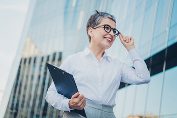 Confident businesswoman with short grey hair holding clipboard in urban setting with modern glass...