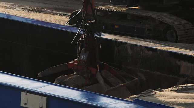 An excavator with a clamshell bucket unloads sand from a cargo barge at an industrial port on a sunny day