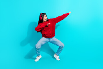 Young woman dancing joyfully in a casual red sweater and jeans wearing headphones on a vibrant turquoise background